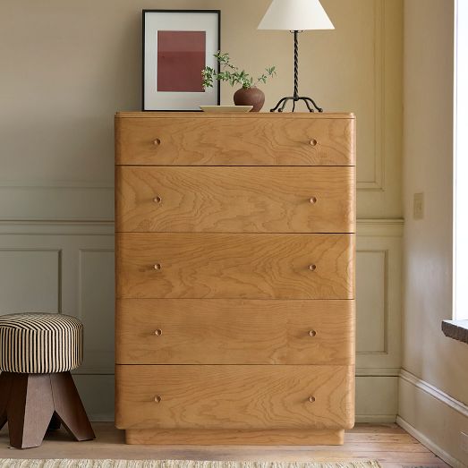 A light wood dresser with six drawers, a striped upholstered stool to the left, and a lamp, framed art, and small plant arranged on top.