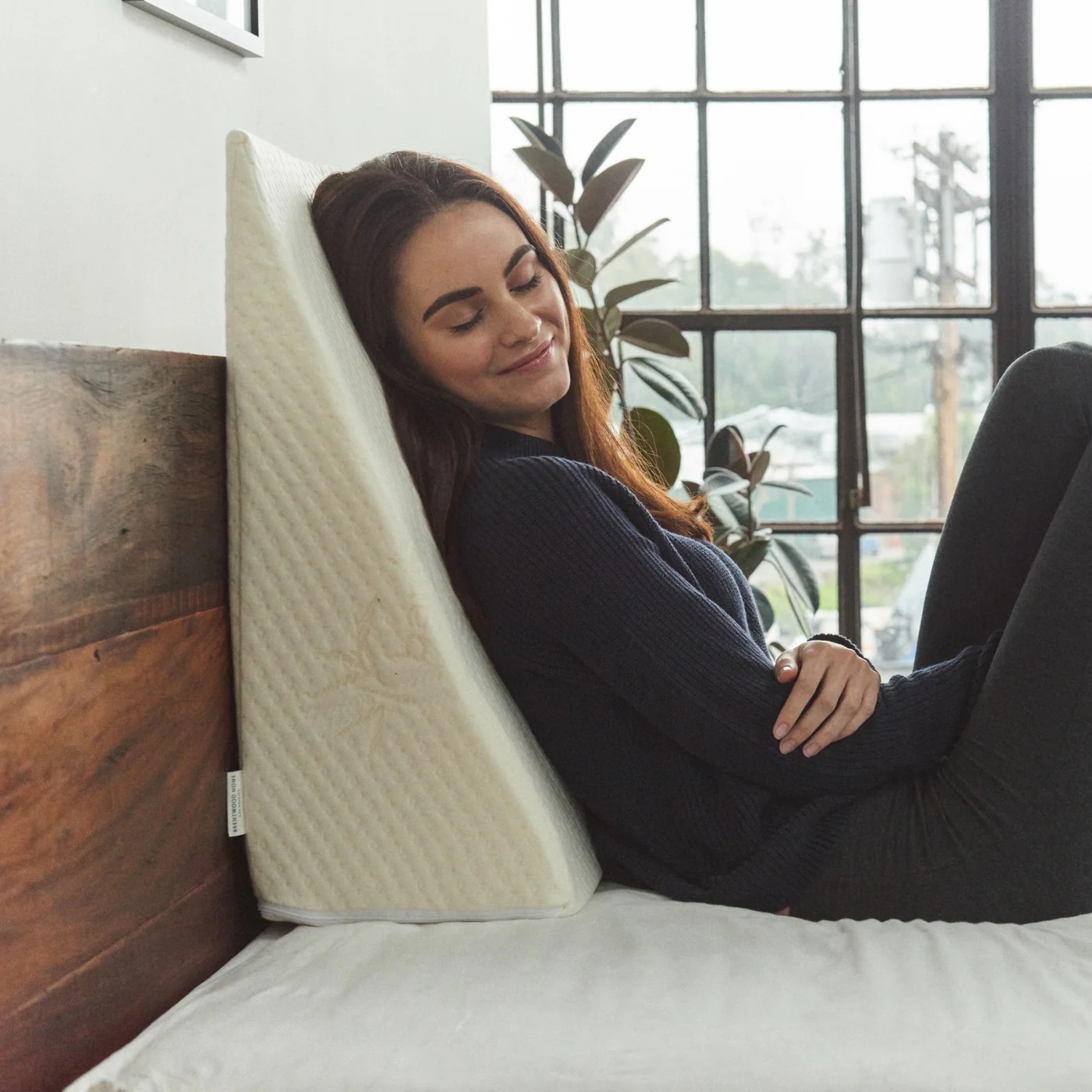 A woman leans comfortably against a large triangular wedge pillow on a bed, with a window and plants in the background.
