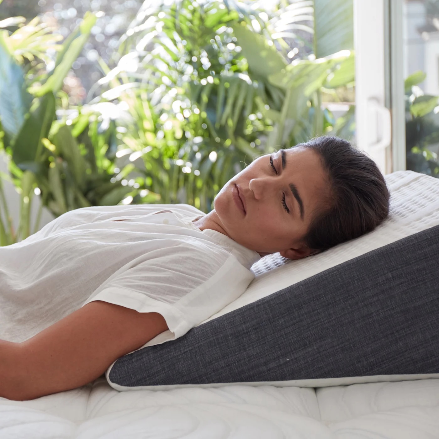 A woman rests on a bed using a wedge pillow, with sunlight and green plants visible through a window in the background.
