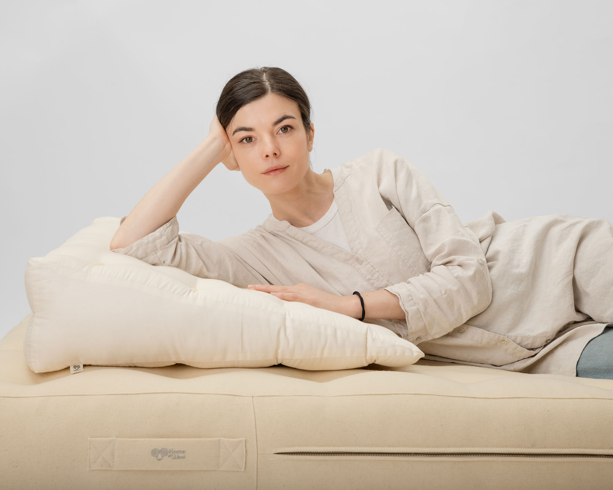 A woman in beige clothing reclines on a cream-colored mattress, propped up on pillows, looking directly at the camera against a plain background.