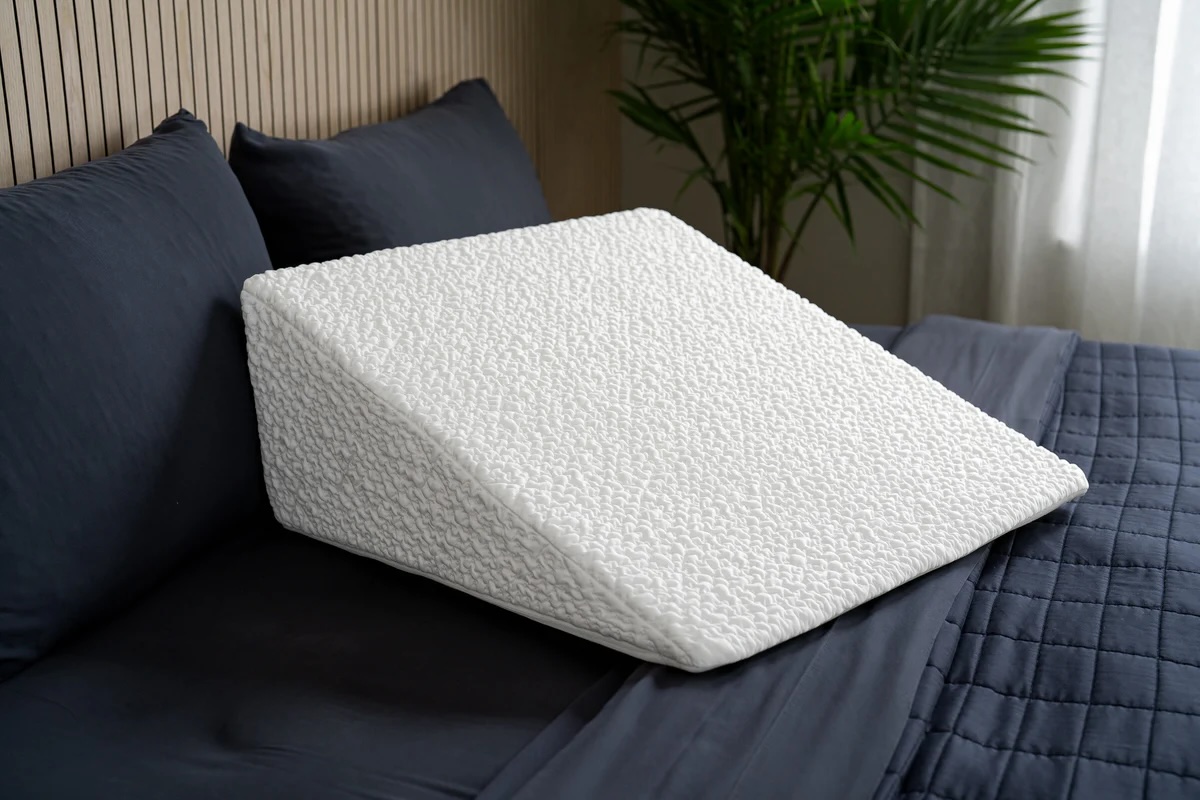 A white wedge pillow with a textured cover is placed on a dark blue bed with matching pillows and duvet, beside a green plant.