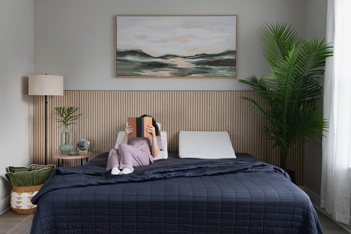 Person lying on a bed with a navy quilt, reading a book. There’s a potted plant, lamp, and wall art in a modern, minimal bedroom.