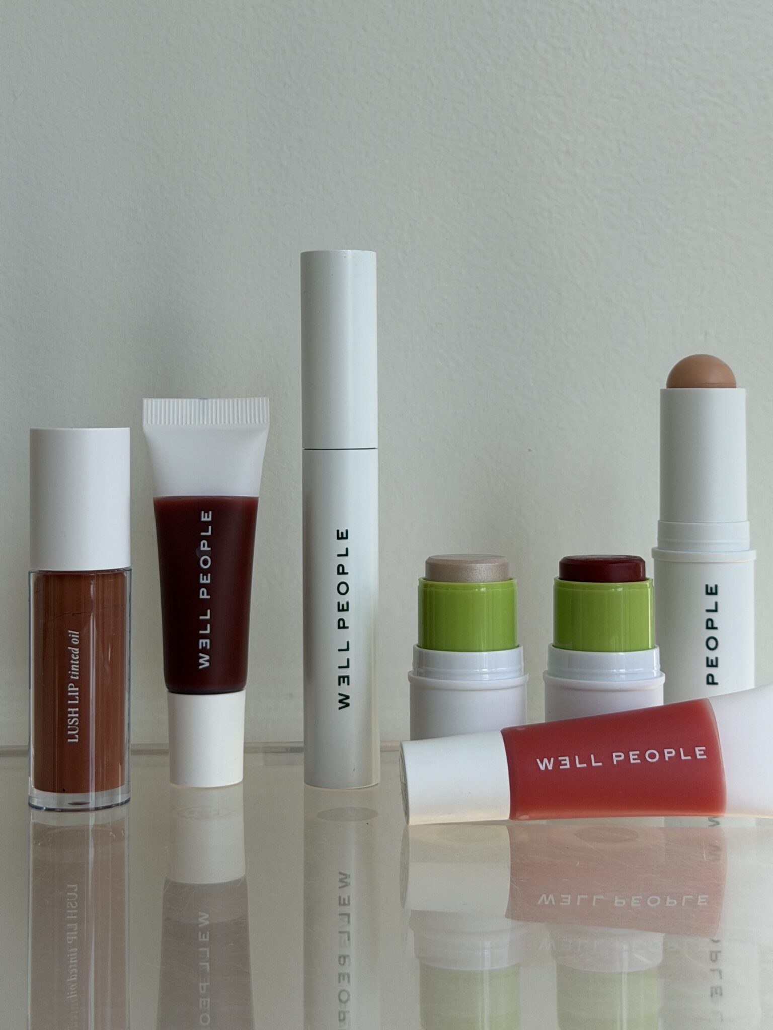 A collection of six Well People makeup products including lip gloss, tubes, and stick applicators, arranged upright and lying down on a reflective surface against a plain background.