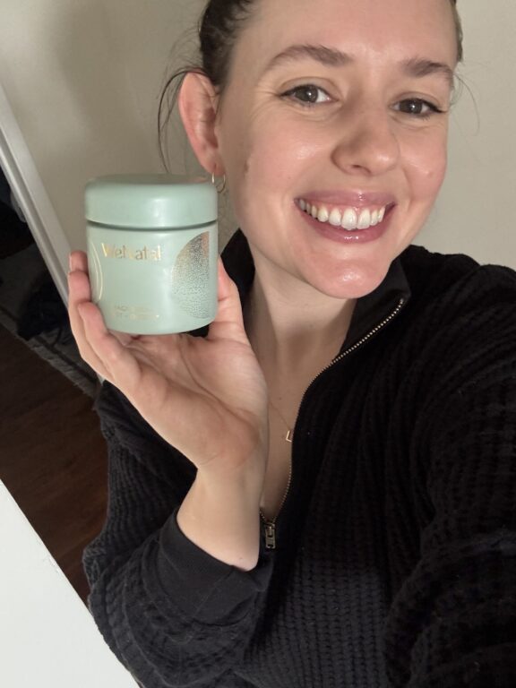 A smiling person in a black zip-up top holds up a green container labeled "Violetal" in their left hand, posing for a selfie indoors.