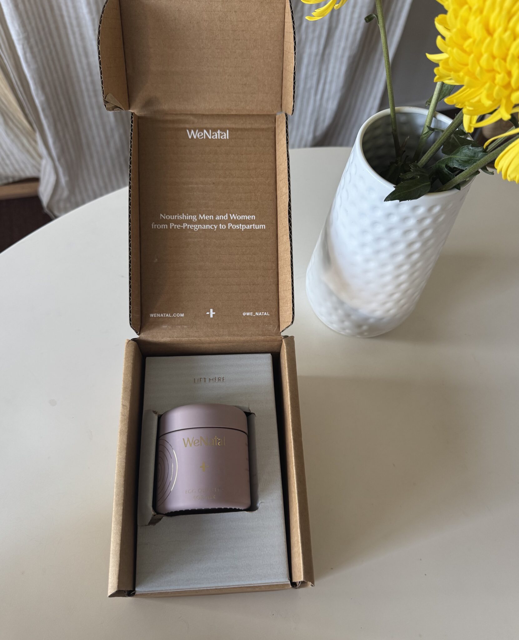 A WeNatal supplement container sits inside an open cardboard box on a table next to a white vase with yellow flowers.