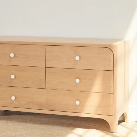 A light wood dresser with six drawers and white round knobs sits against a white wall in natural sunlight.