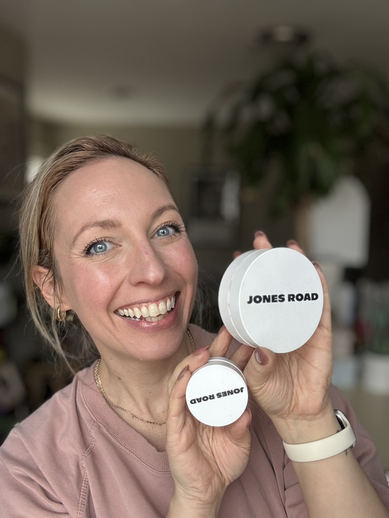 A woman smiling and holding two white cosmetic containers labeled "JONES ROAD" in a well-lit room, reminiscent of Ilia's minimalistic beauty aesthetic.