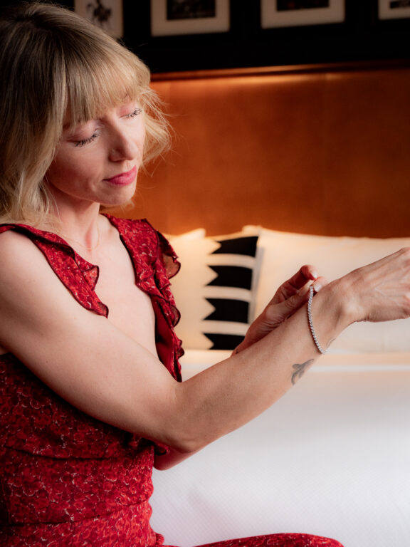 A woman in a red dress sits on a bed and fastens a bracelet on her wrist, with a pillow and framed pictures in the background.