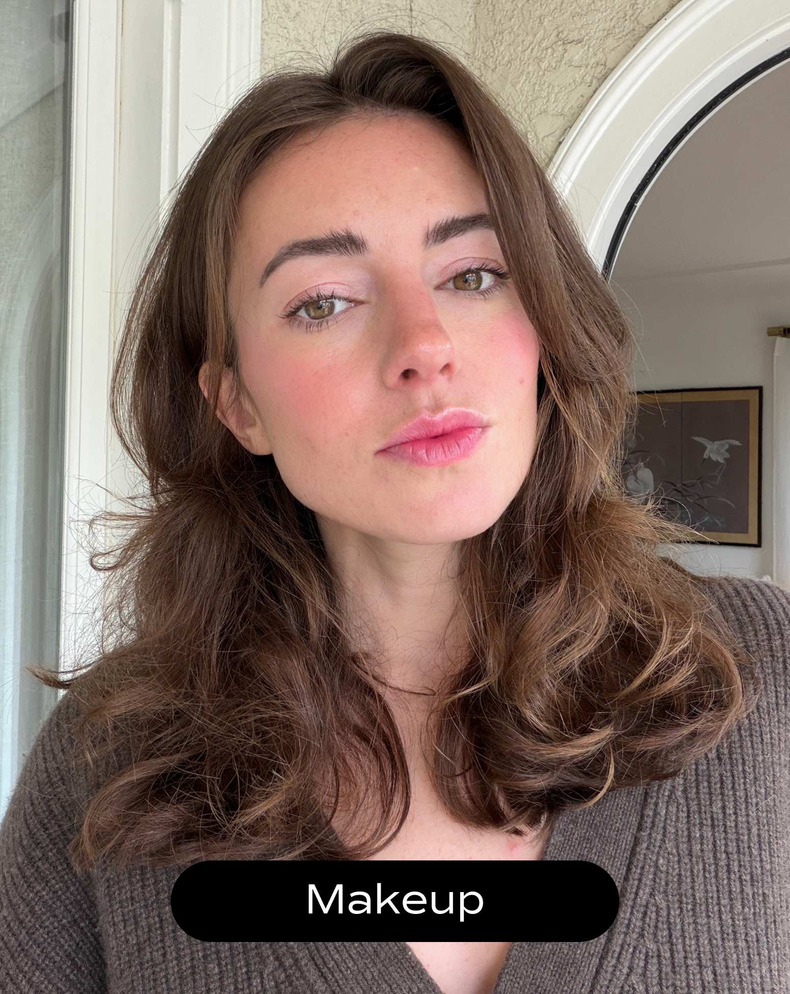 Woman with light skin and brown hair poses indoors, wearing subtle makeup and a brown sweater. A framed picture is visible in the background. The text "Makeup" appears at the bottom.