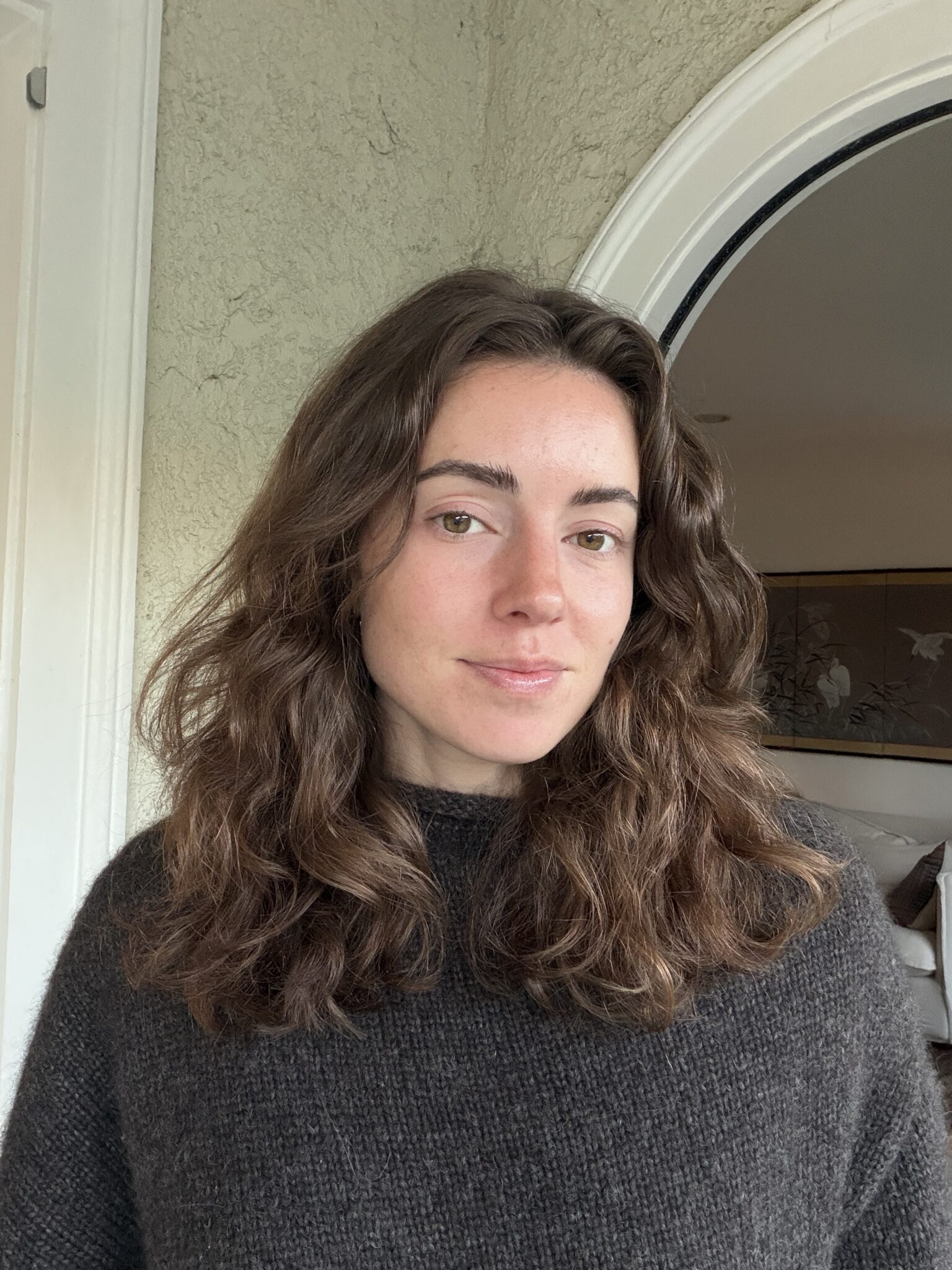 A woman with wavy brown hair and a dark sweater stands indoors in front of a textured wall and an arched doorway.