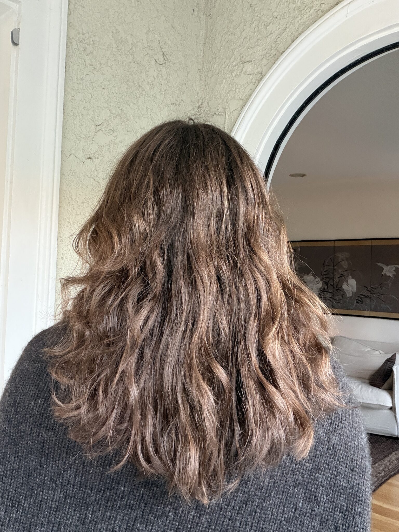 Person with medium-length, wavy brown hair seen from behind, standing indoors near an arched doorway with partial view of a living room.