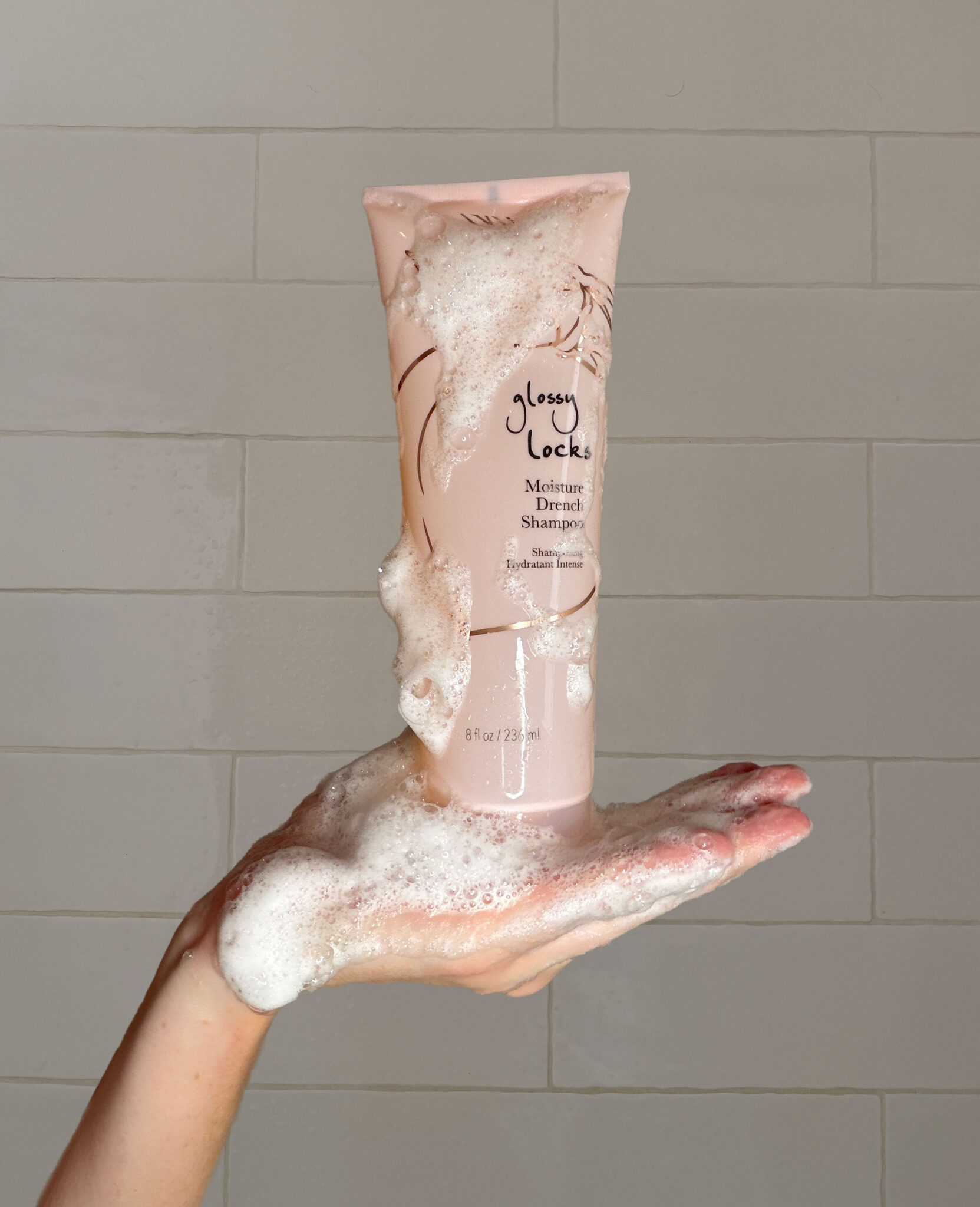 A hand holds a pink bottle of Glossy Locks shampoo covered in white foamy bubbles against a tiled background.