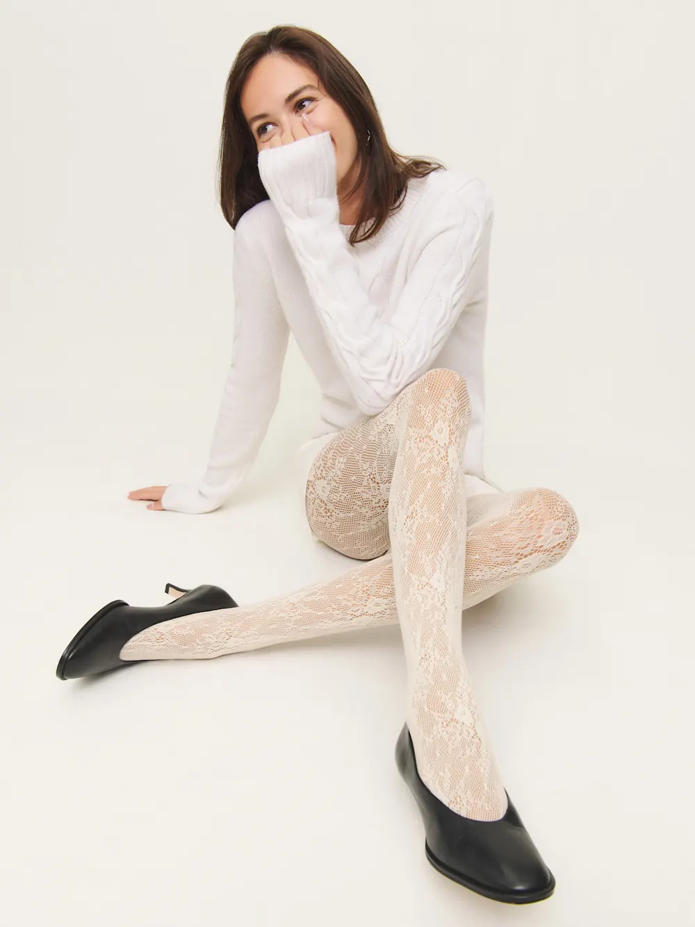 A woman sits on the floor wearing a white sweater, white lace tights, and black flats, with one hand covering her mouth and her legs crossed.
