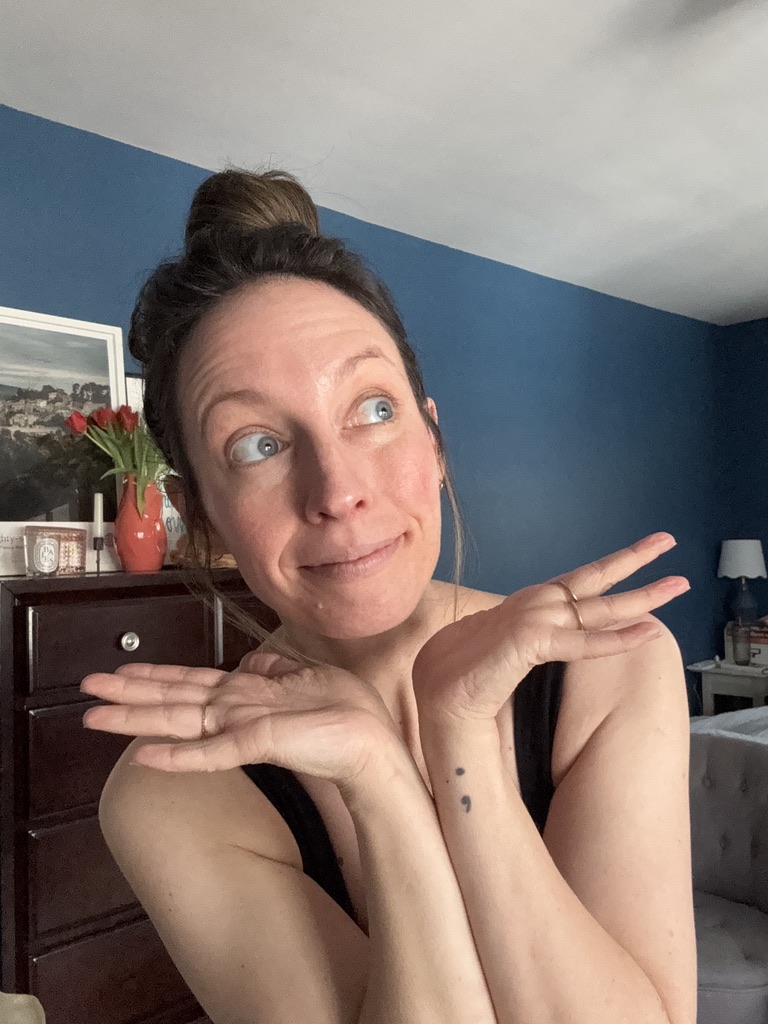 A woman with a topknot hairstyle shrugs with both hands raised, standing in a blue-walled room featuring a dresser, flowers, a framed picture, and Ilia decor accents in the background.