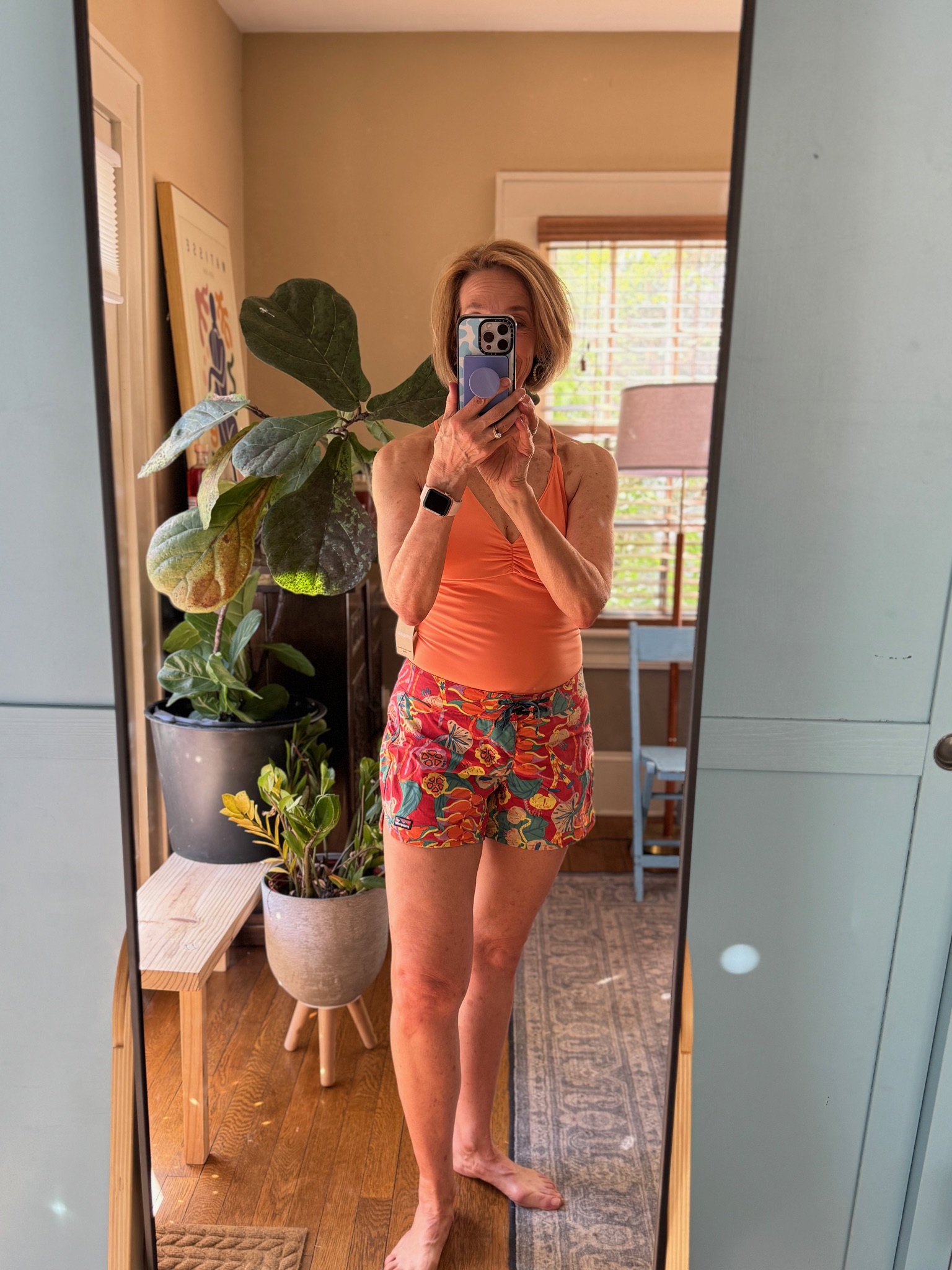 A person stands barefoot in front of a mirror, wearing an orange top and colorful shorts, holding a phone to take a selfie in a sunlit room with indoor plants and wooden furniture.
