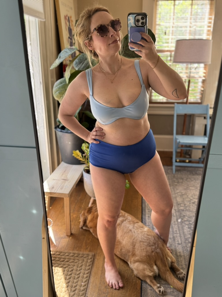 Woman wearing sunglasses and a blue bikini takes a mirror selfie in a sunlit room. A dog lies on the floor near her feet.