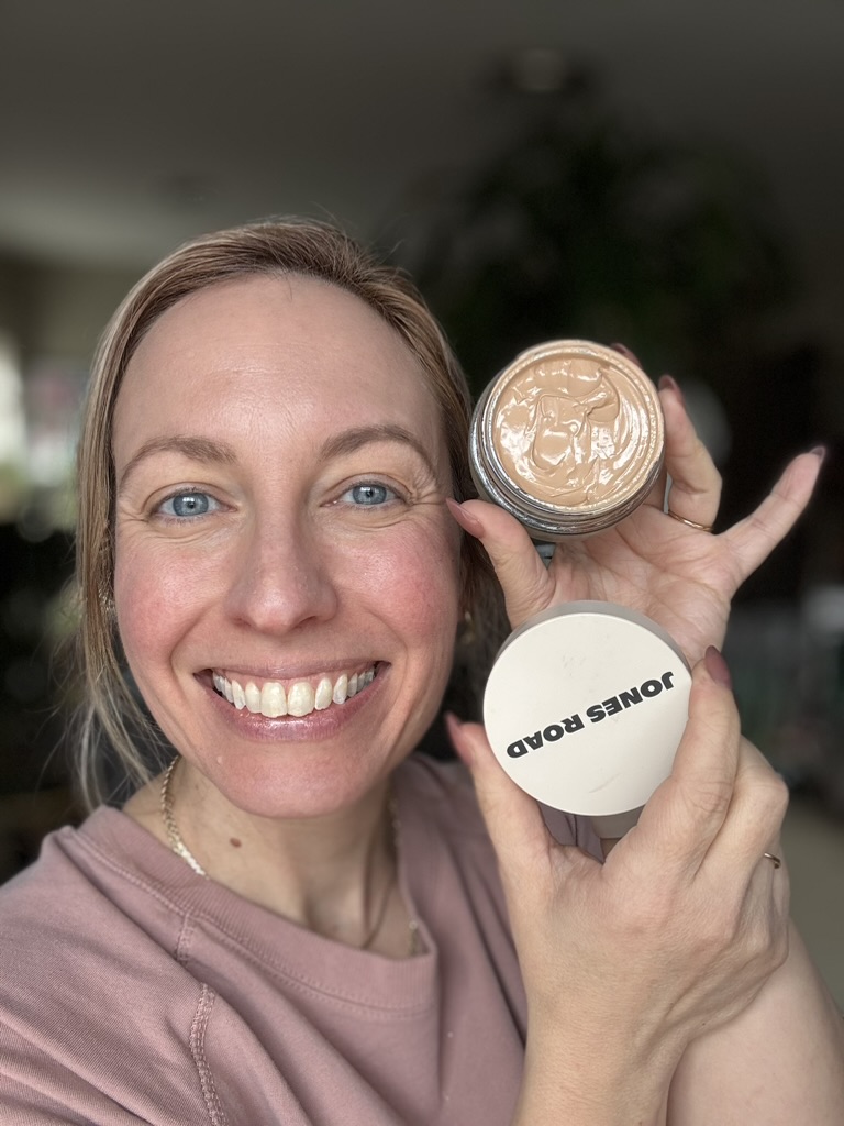 Smiling woman holds an open jar of Jones Road makeup foundation in one hand and the product lid in the other, showing both to the camera—her Ilia-inspired look glowing with natural radiance.
