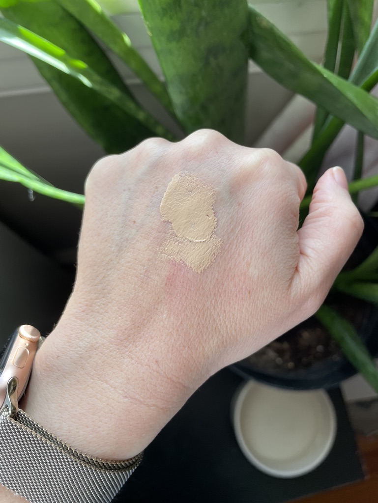 A hand with two swatches of Ilia beige makeup on the back, positioned in front of a green potted plant.