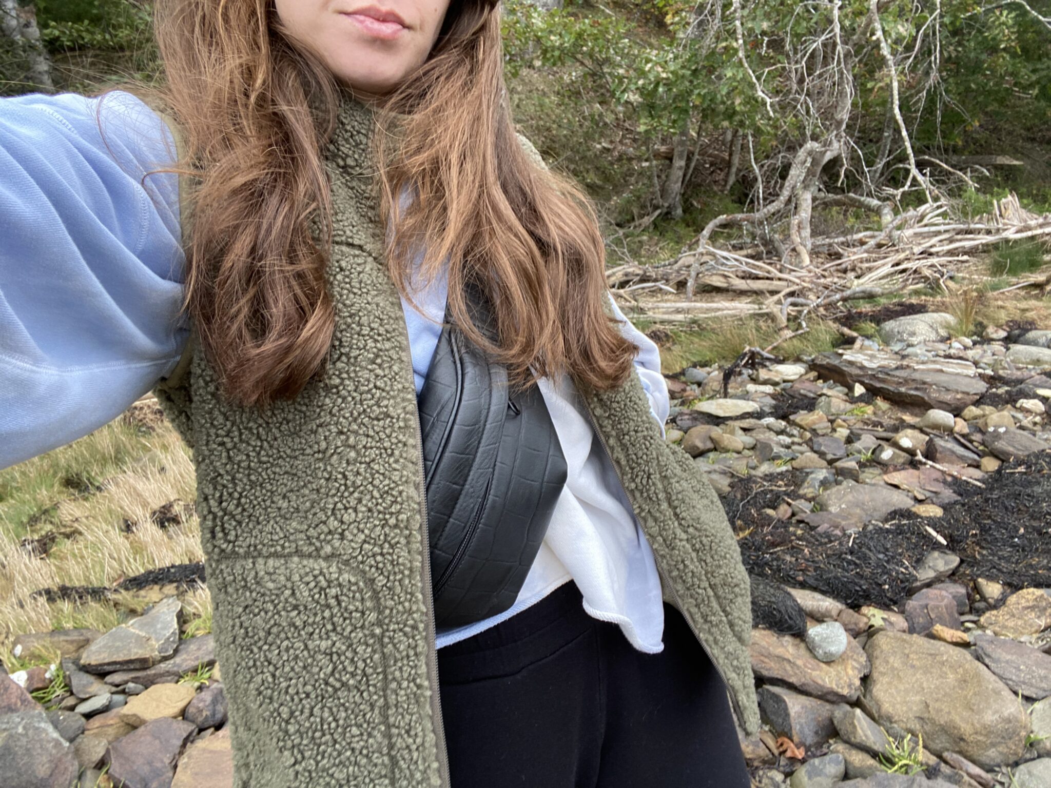 A person with long brown hair wears a green fleece vest, blue sweatshirt, and crossbody bag, standing outdoors on rocky ground with plants and driftwood in the background.