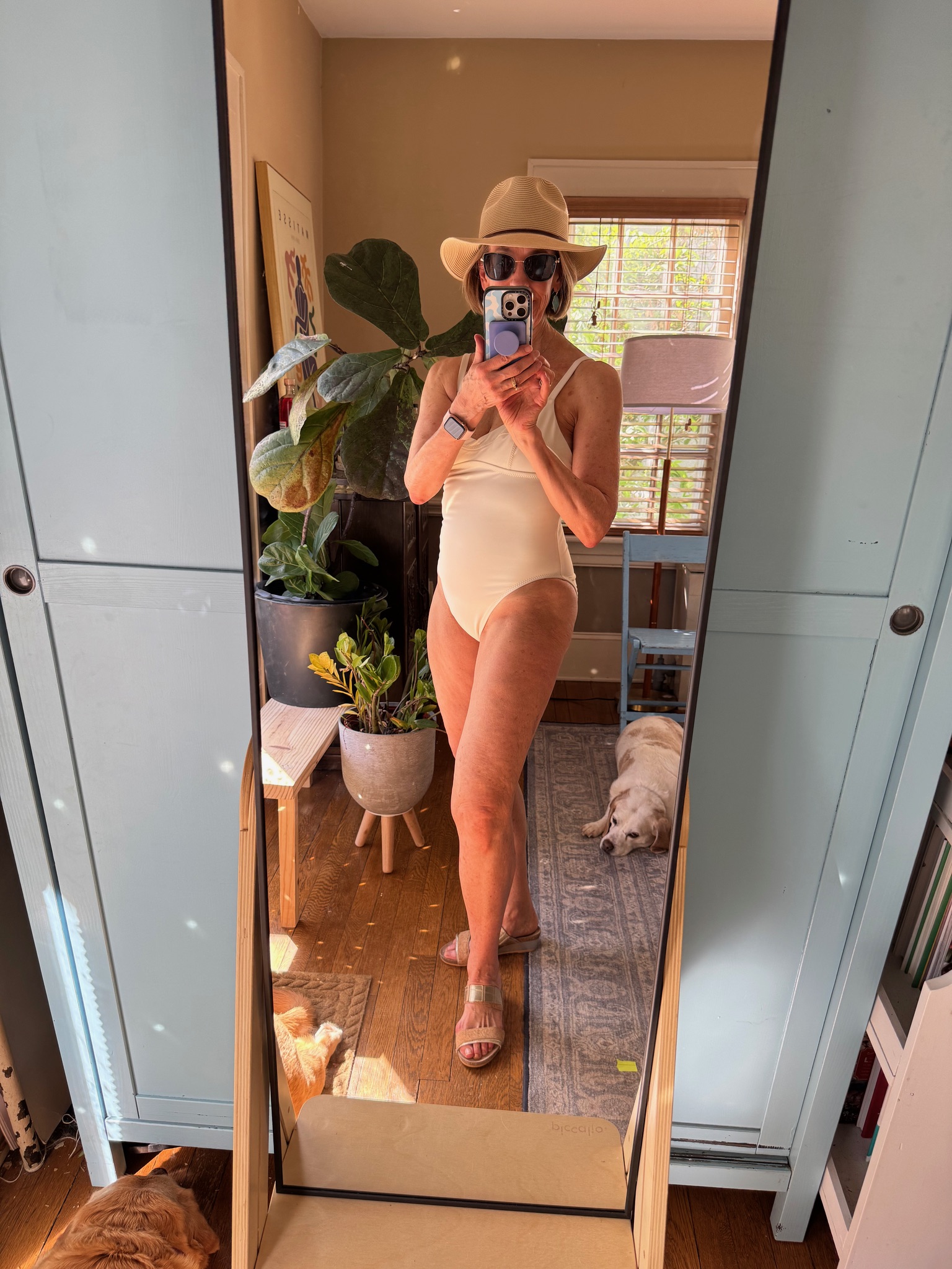 Person in a light swimsuit, hat, and sunglasses takes a mirror selfie in a room with plants and a dog lying on the floor.