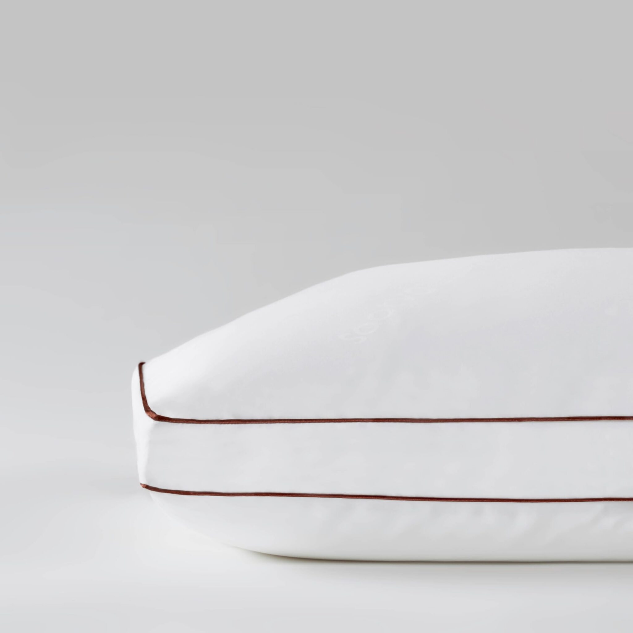 A close-up of a white pillow with brown piping on the edges against a plain light gray background.