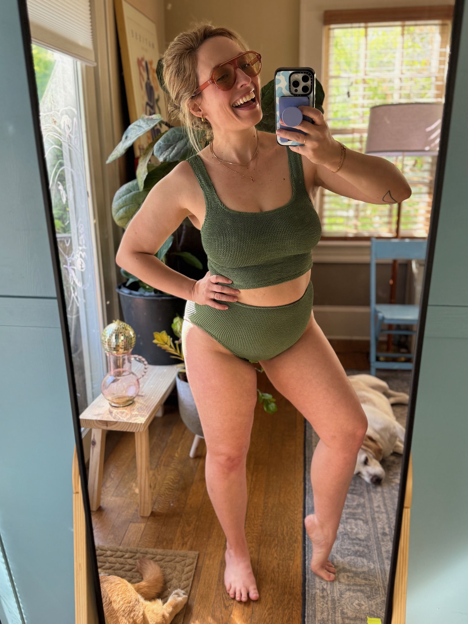 Woman in a green two-piece outfit takes a smiling mirror selfie in a sunlit room with plants, a lamp, and two dogs lying on the floor.