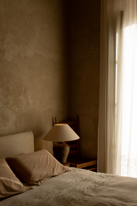 A softly lit bedroom with a beige bed, pillows, a bedside table, a ceramic lamp, a wooden chair, and sheer curtains covering a window.