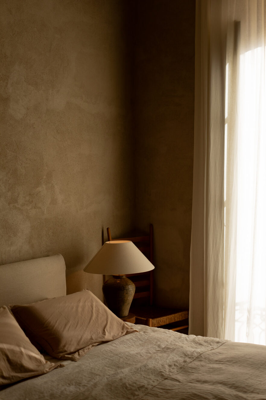 A softly lit bedroom with a beige bed, pillows, a bedside table, a ceramic lamp, a wooden chair, and sheer curtains covering a window.