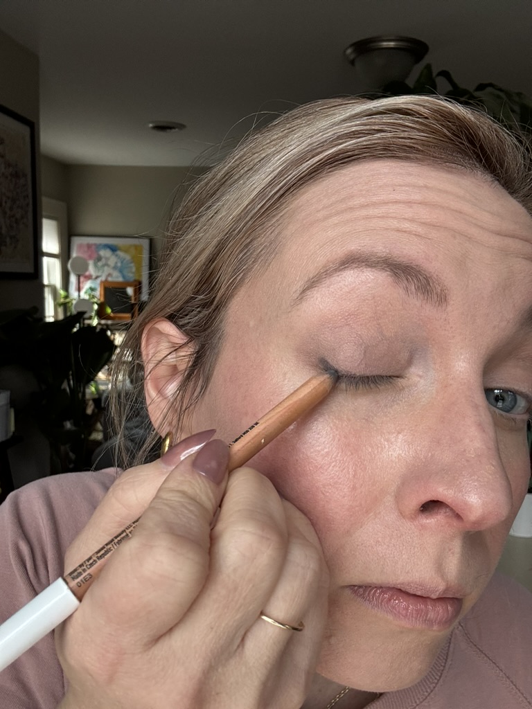 A person applies Ilia makeup with a pencil to their upper eyelid, holding their eyelid taut while looking slightly downward.