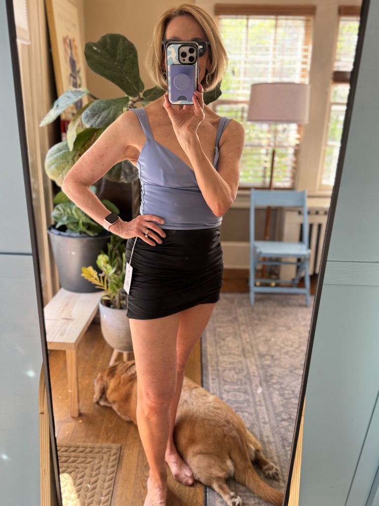A woman takes a mirror selfie wearing a lavender top and black skirt. A dog lies on the floor behind her in a well-lit room with plants and furniture.