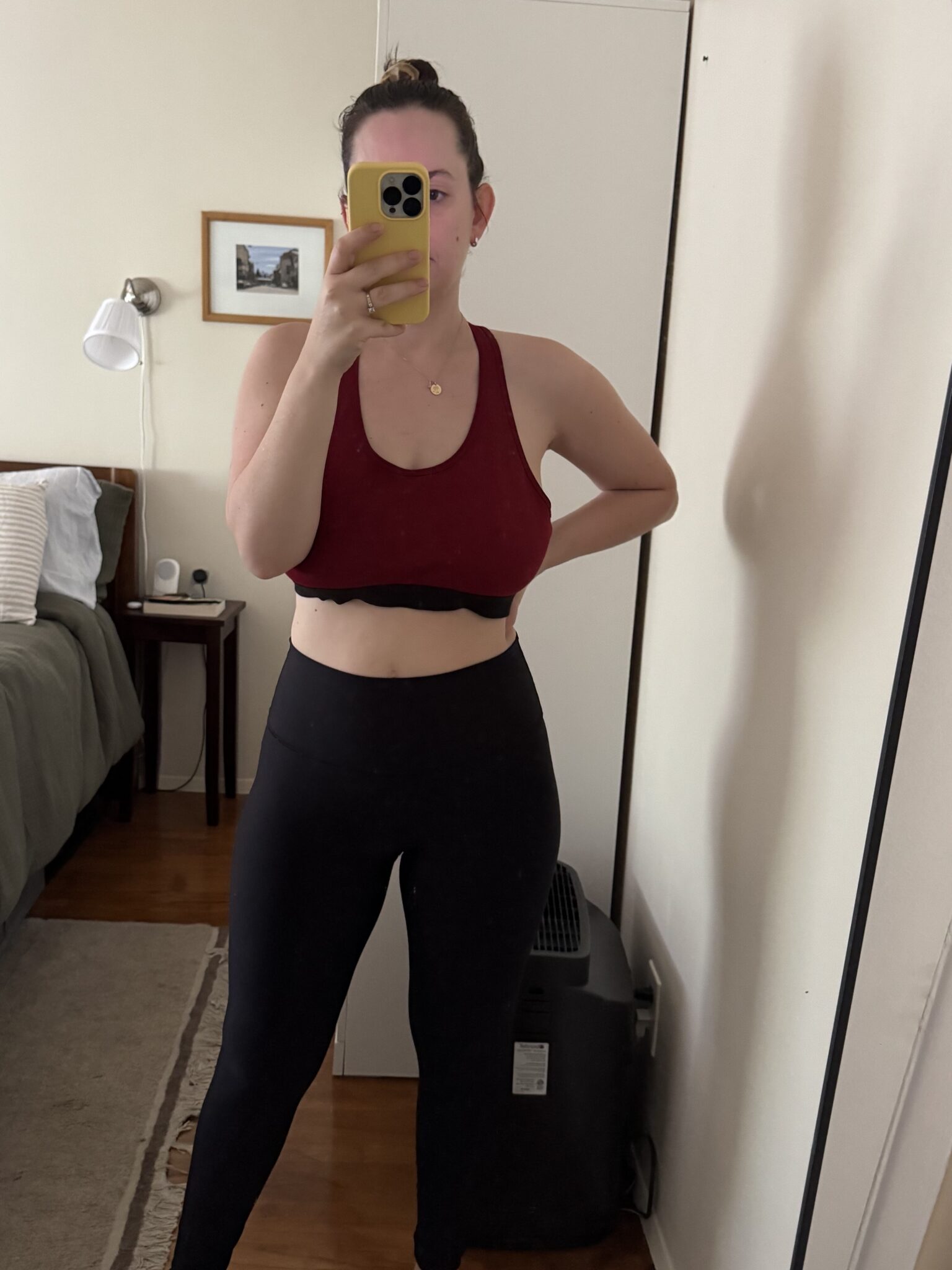 Person wearing a red sports bra and black leggings taking a mirror selfie in a bedroom with a bed, nightstand, and basket on a wardrobe.