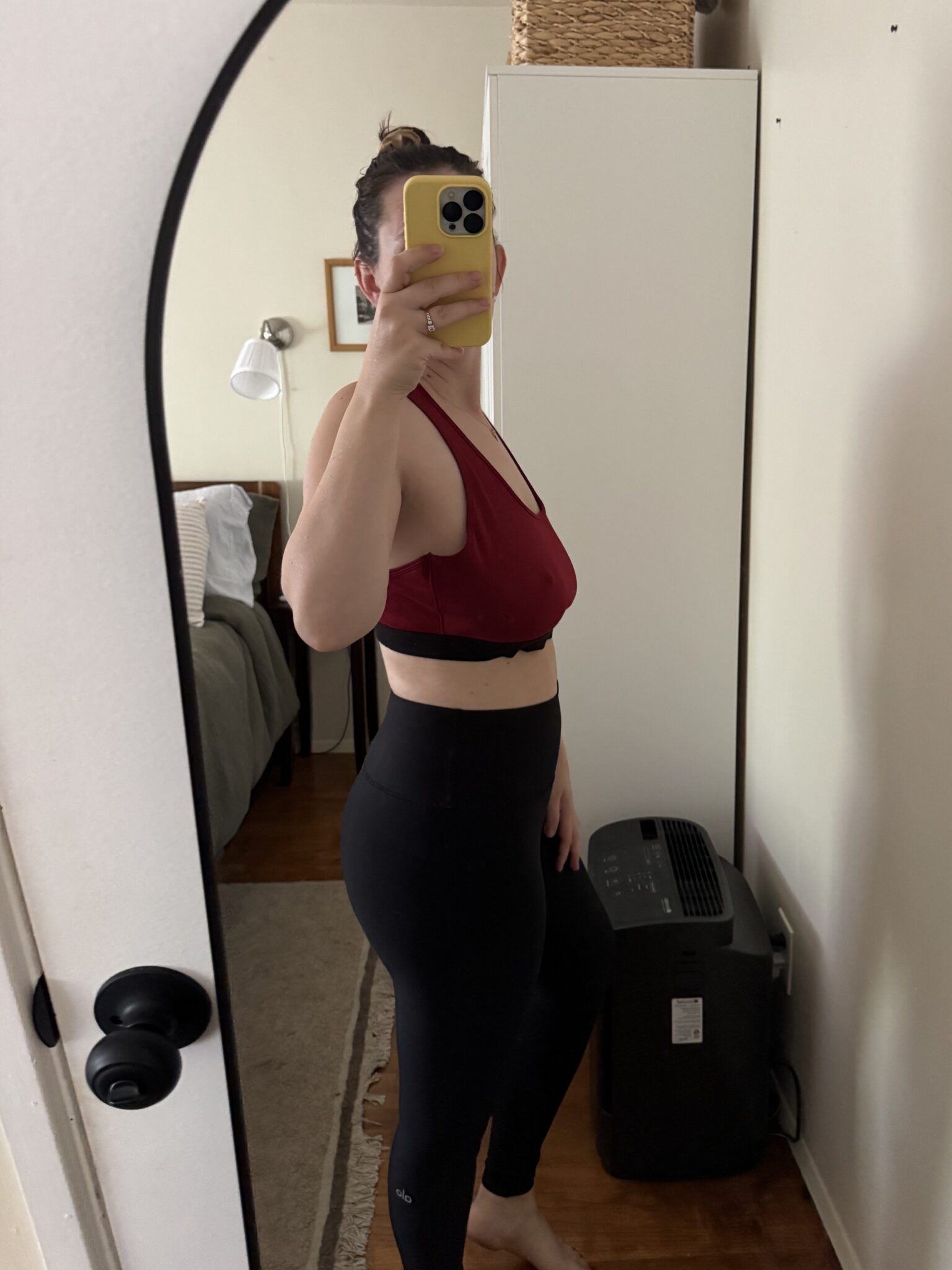 A person stands sideways in front of a mirror, wearing a red sports bra and black leggings, holding a phone to take a mirror selfie in a bedroom.