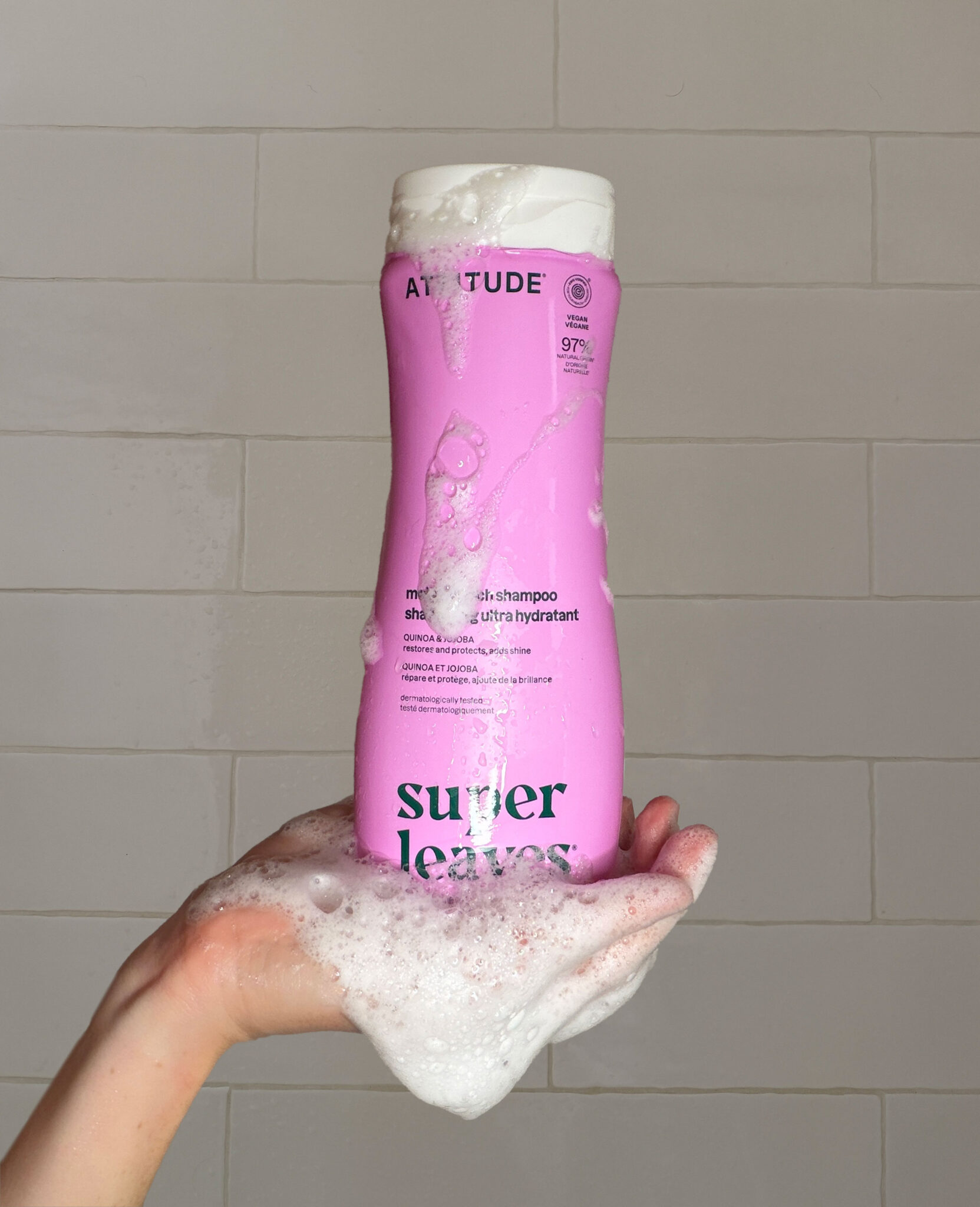 A hand holds a pink bottle of Attitude Super Leaves shampoo covered in soap suds against a tiled background.