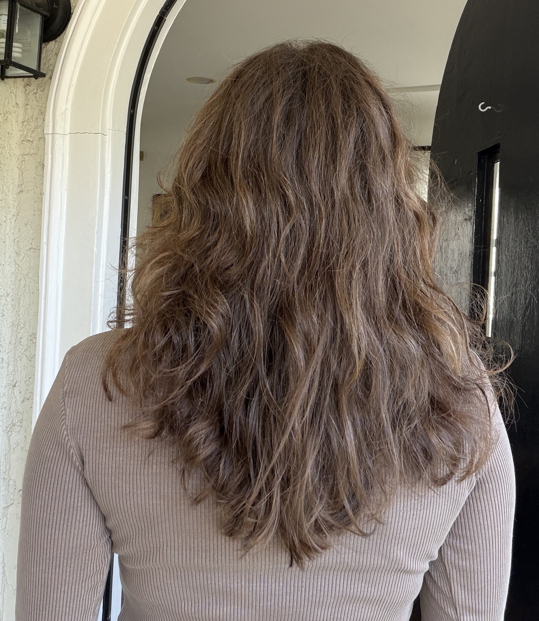 A person with long, wavy brown hair is standing with their back to the camera in a doorway, wearing a ribbed, light brown top.