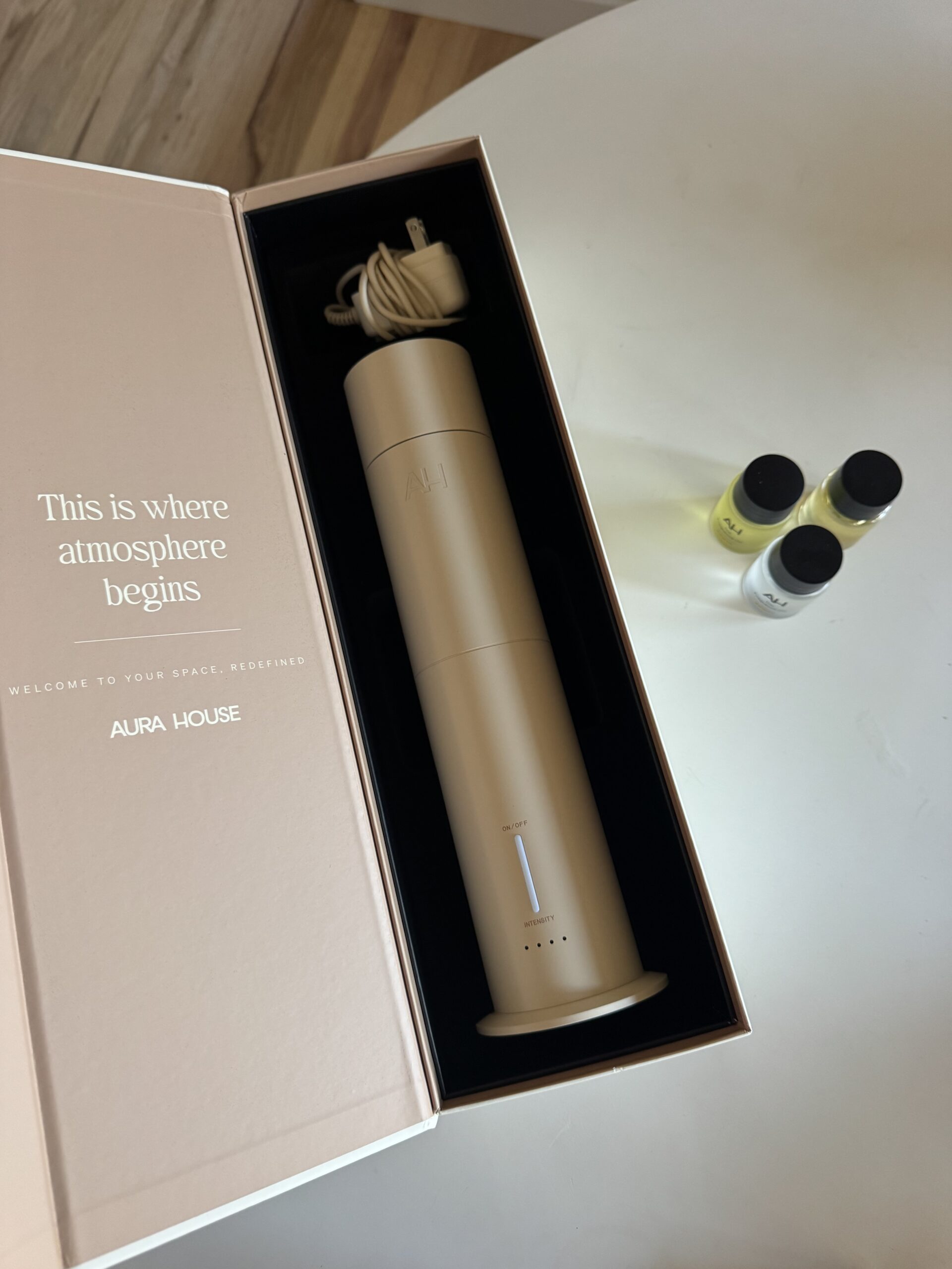 A gold cylindrical diffuser in a box with “This is where atmosphere begins” text, next to three small bottles of fragrance oil on a white surface.