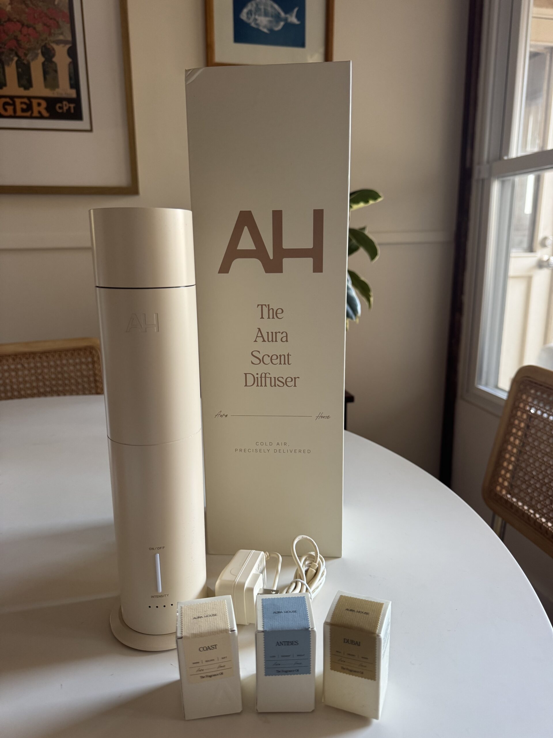 A white scent diffuser with its box is on a table, next to three scent capsules labeled Coast, Artemis, and Balm, in a room with art on the walls and a window.