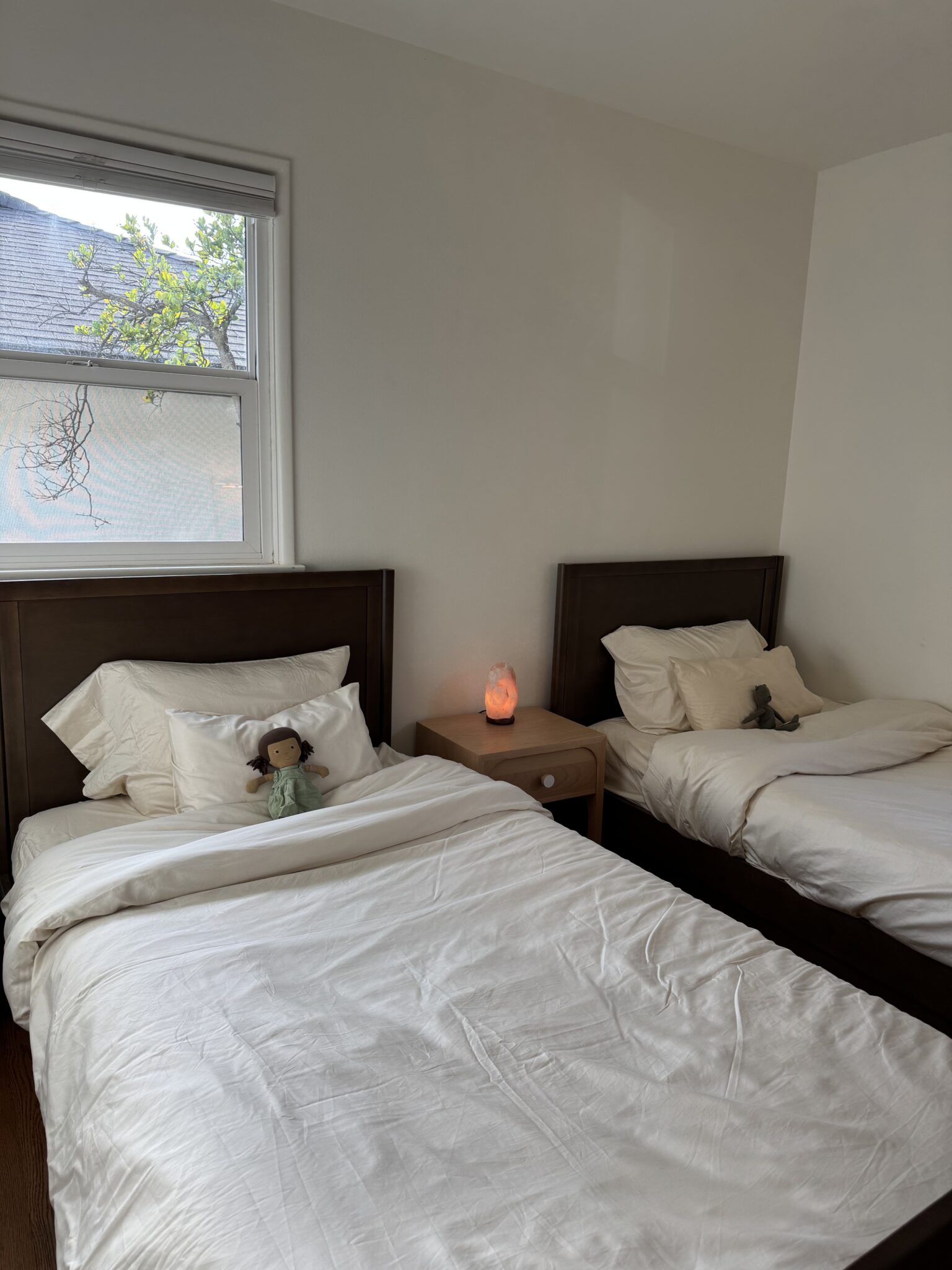 Two single beds with white bedding are placed in a corner of a room. A nightstand with a salt lamp sits between them. Each bed has a doll or stuffed animal on the pillow.