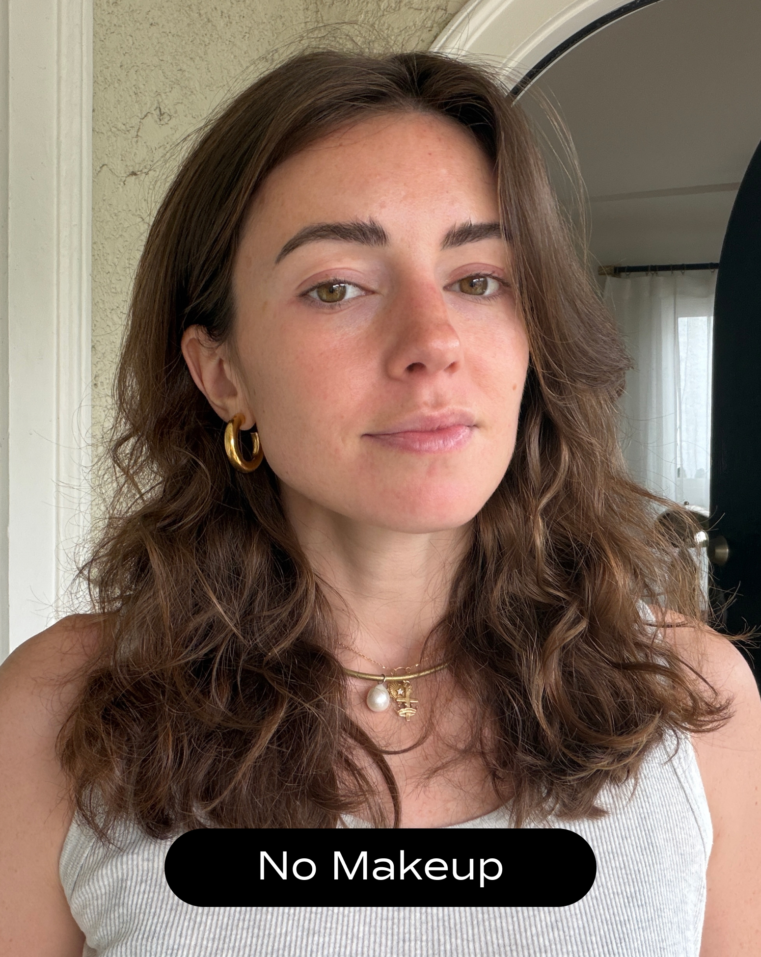 A woman with wavy brown hair and gold jewelry stands indoors, wearing a white tank top. A label at the bottom reads "No Makeup.