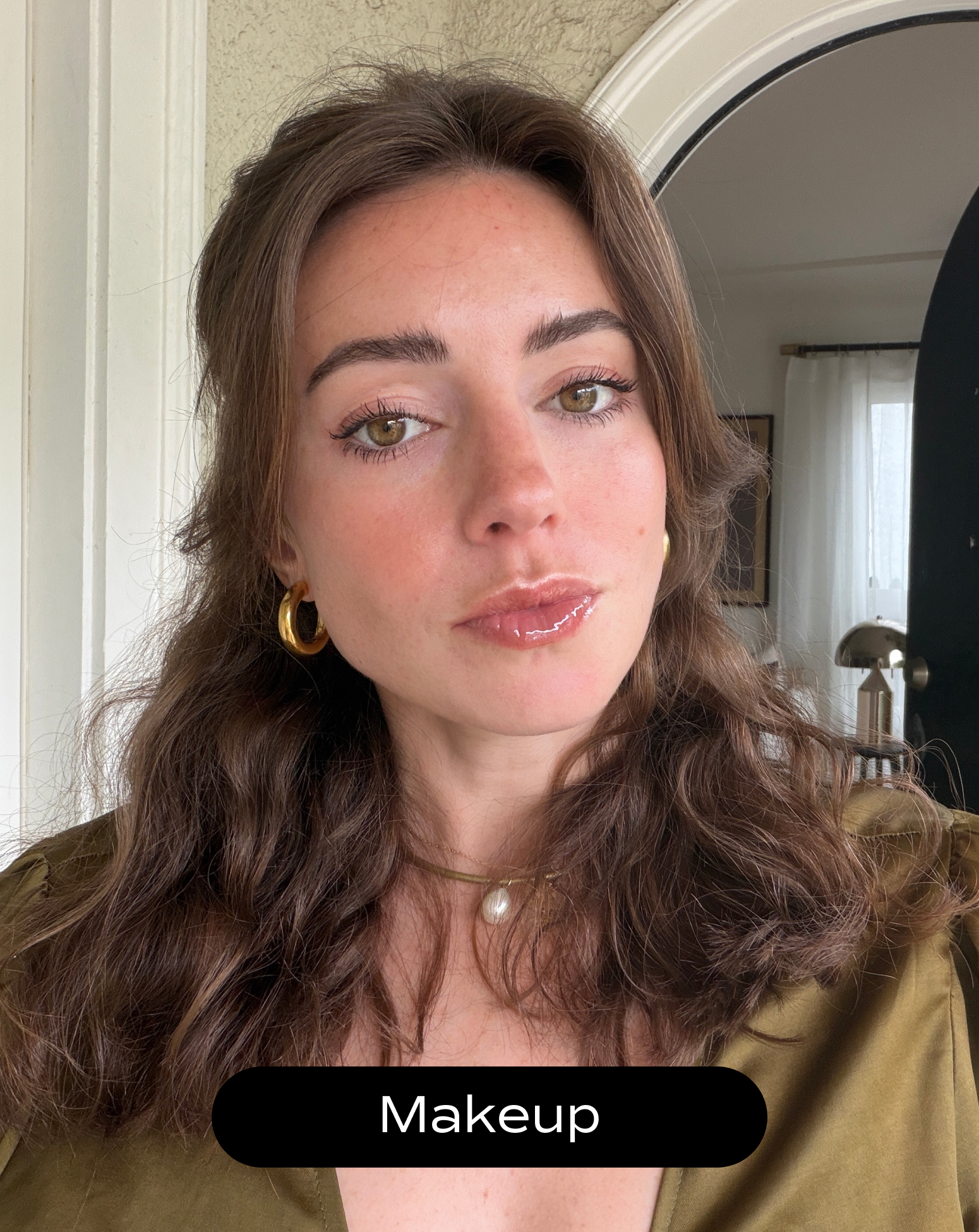 Woman with wavy brown hair wearing gold hoop earrings, a pearl necklace, and natural makeup; standing indoors near a window and lamp.
