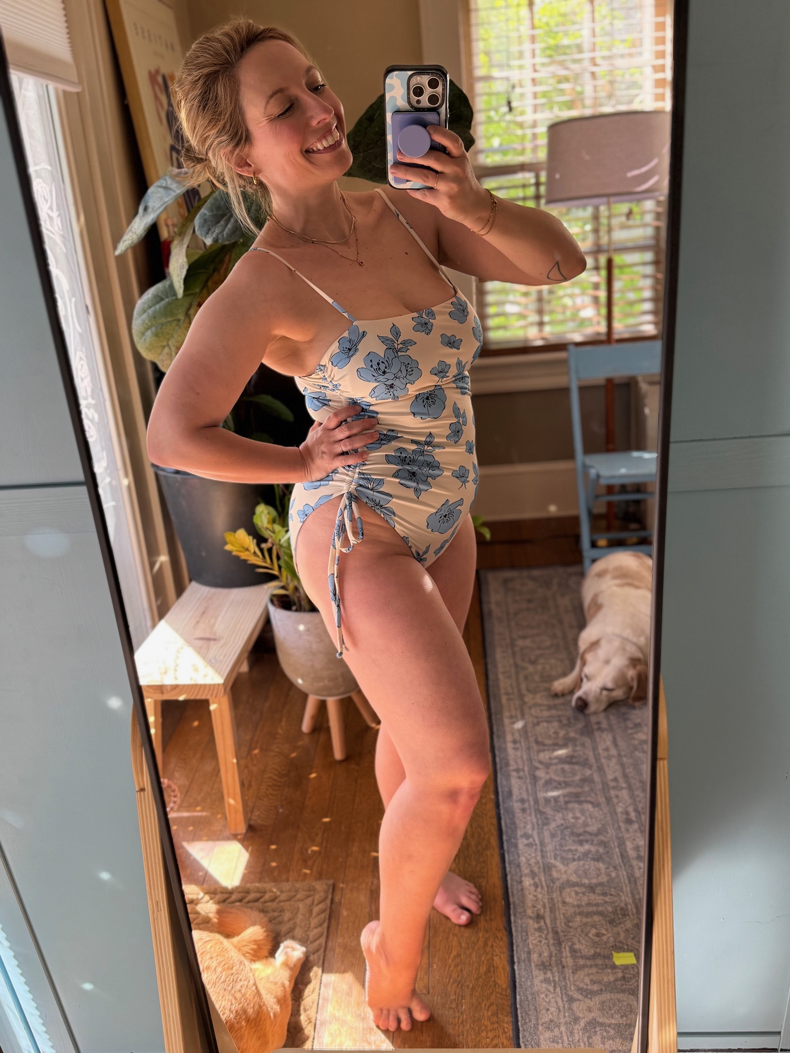 Woman in a floral one-piece swimsuit takes a mirror selfie in a sunlit room with plants, furniture, and a dog lying on the floor in the background.
