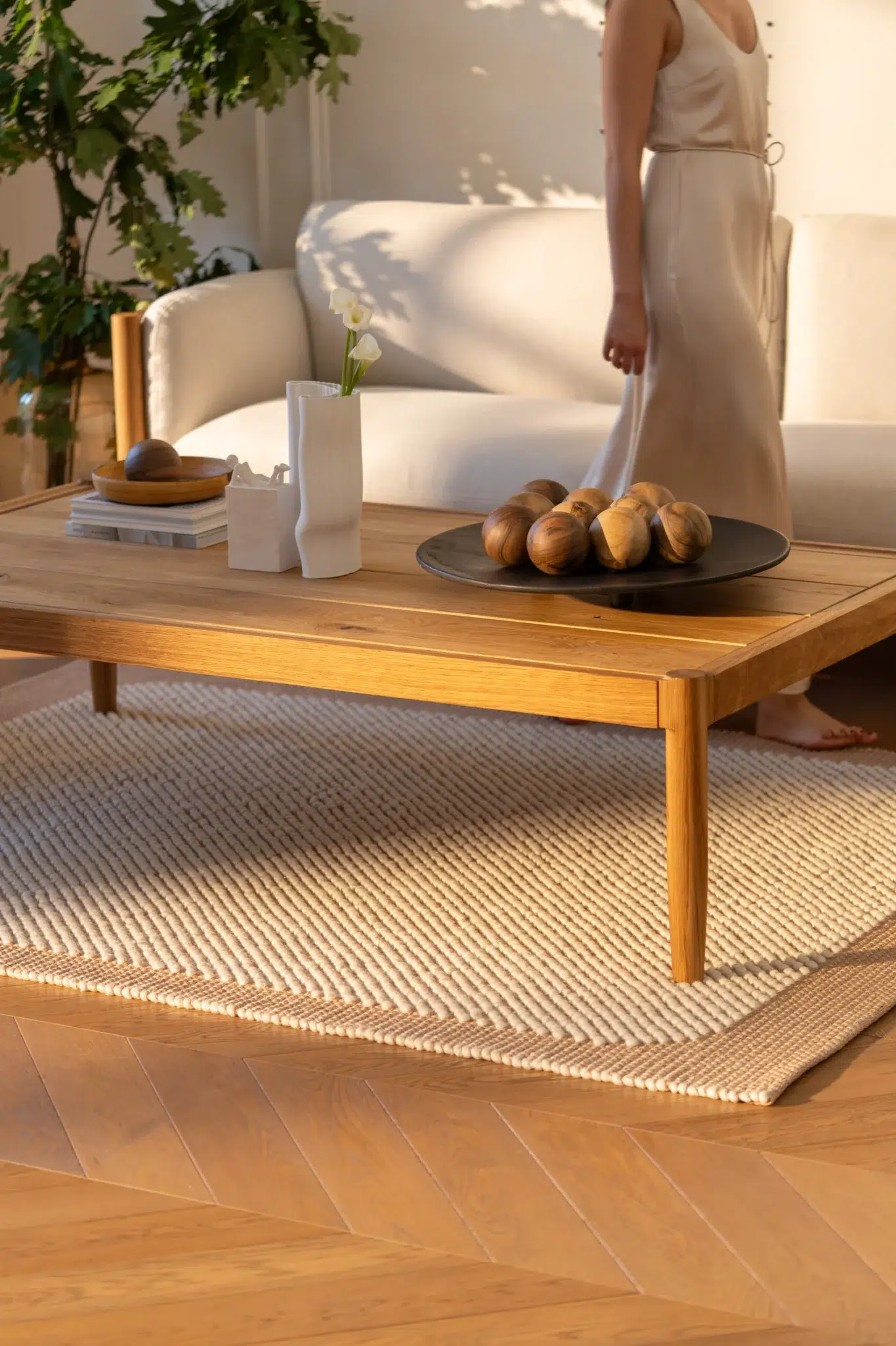A woman in a light dress stands near a beige sofa behind a wooden coffee table with decor items in a sunlit living room.