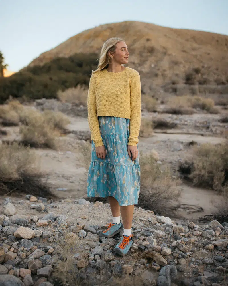 A woman stands outdoors on rocky ground, wearing a yellow knit sweater, a blue patterned skirt, and sneakers, with dry shrubs and a hill in the background.