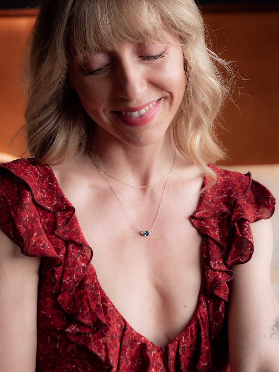A woman with blonde hair in a red ruffled dress smiles with her eyes closed, wearing a delicate necklace with a small pendant.