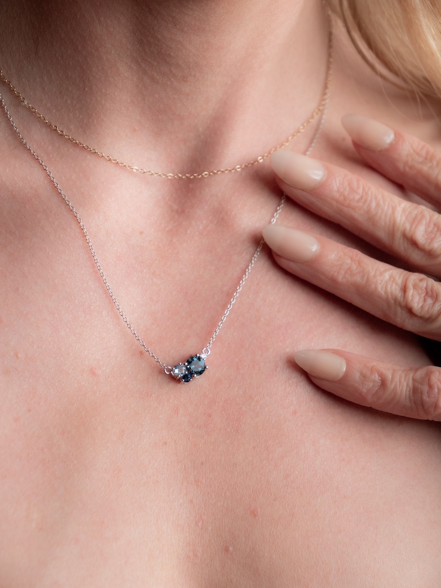 Close-up of a person's neck wearing two delicate necklaces, one with a small cluster of blue gemstones, and a hand with manicured nails resting on their collarbone.
