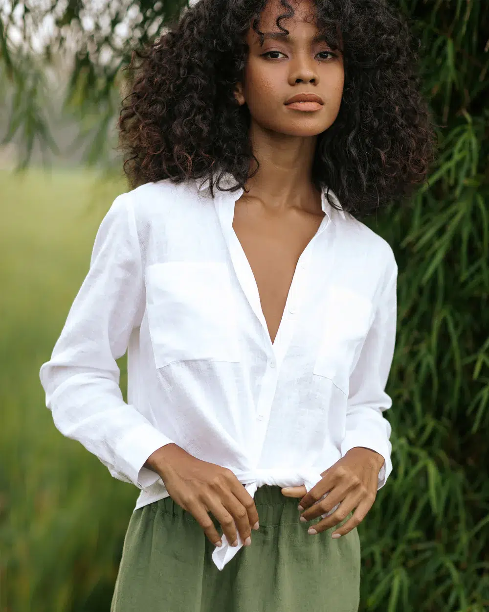 Woman with curly hair wearing a white button-up shirt and green pants stands outdoors in front of greenery.