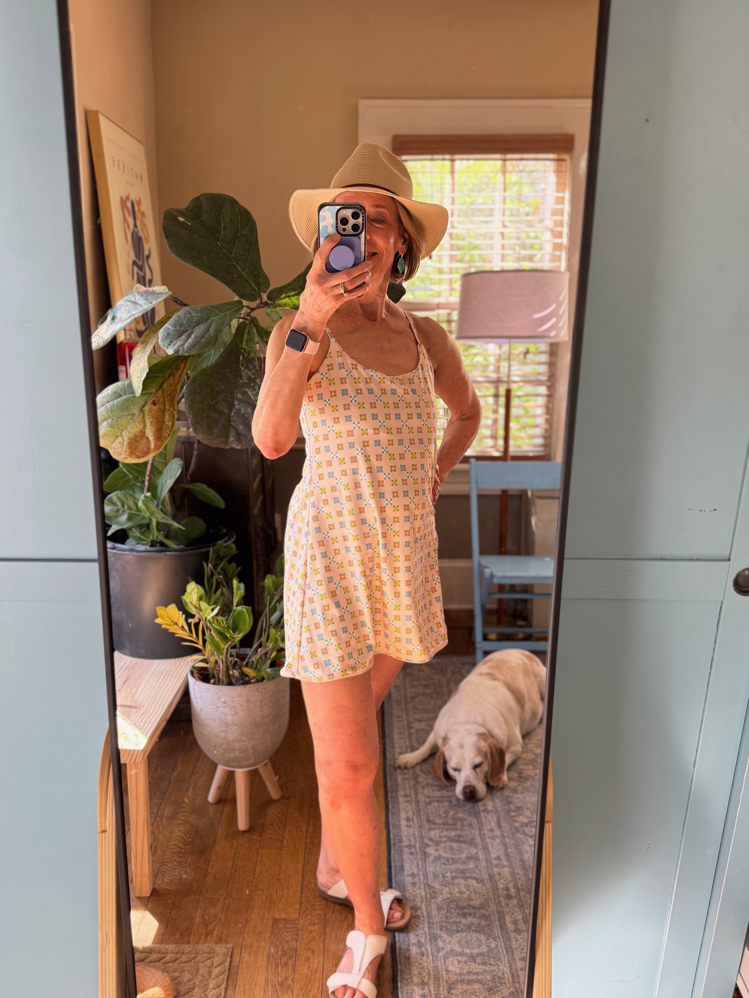 Person wearing a hat and patterned romper takes a mirror selfie in a room with plants and a dog lying on the floor.