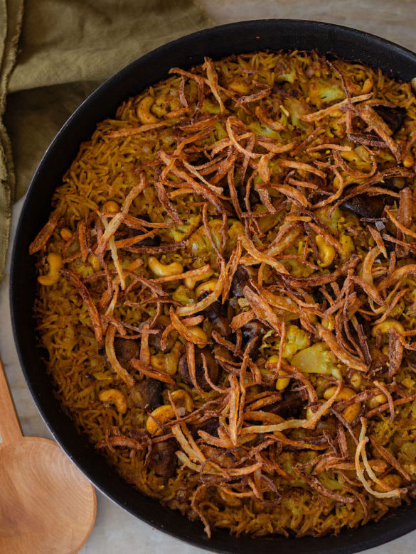 A pan of biryani topped with fried onions, cashews, and spices sits on a marble surface next to a wooden spoon and an olive green cloth napkin.
