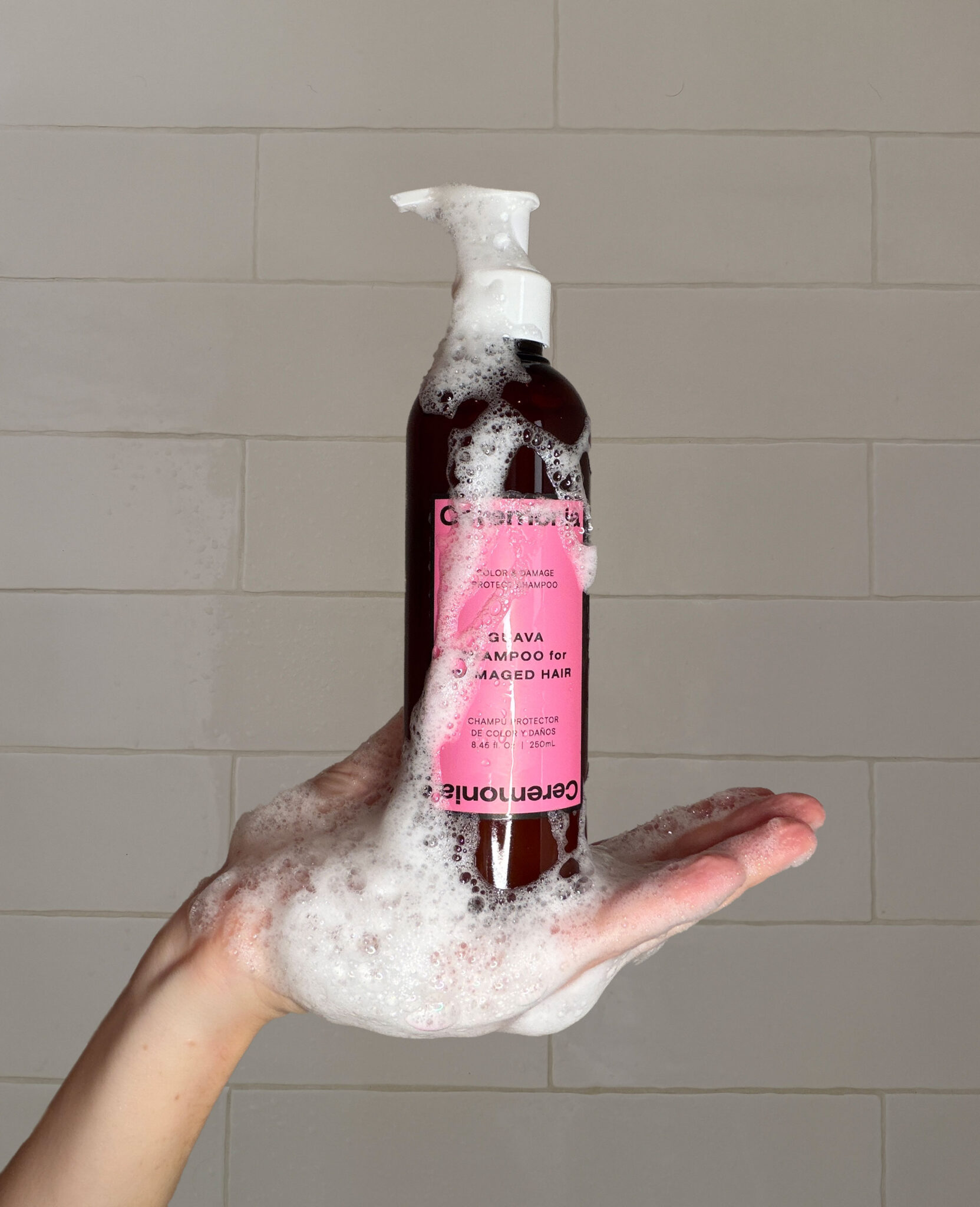 A hand holds a bottle of shampoo covered in foam, with shampoo suds dripping down, against a tiled bathroom wall background.