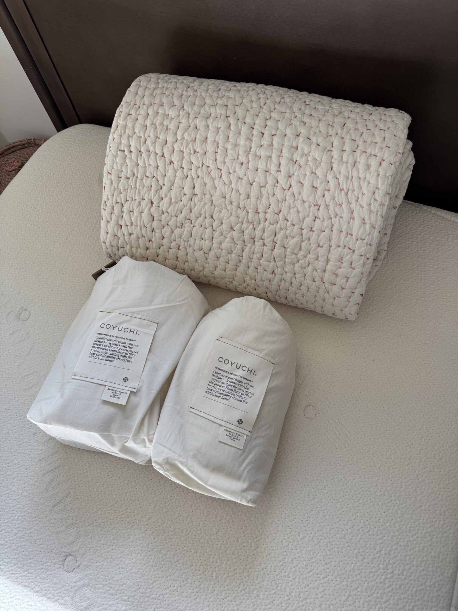 A folded white textured blanket and two packaged Coyuchi bedding items are placed on a mattress with a dark wooden headboard in the background.