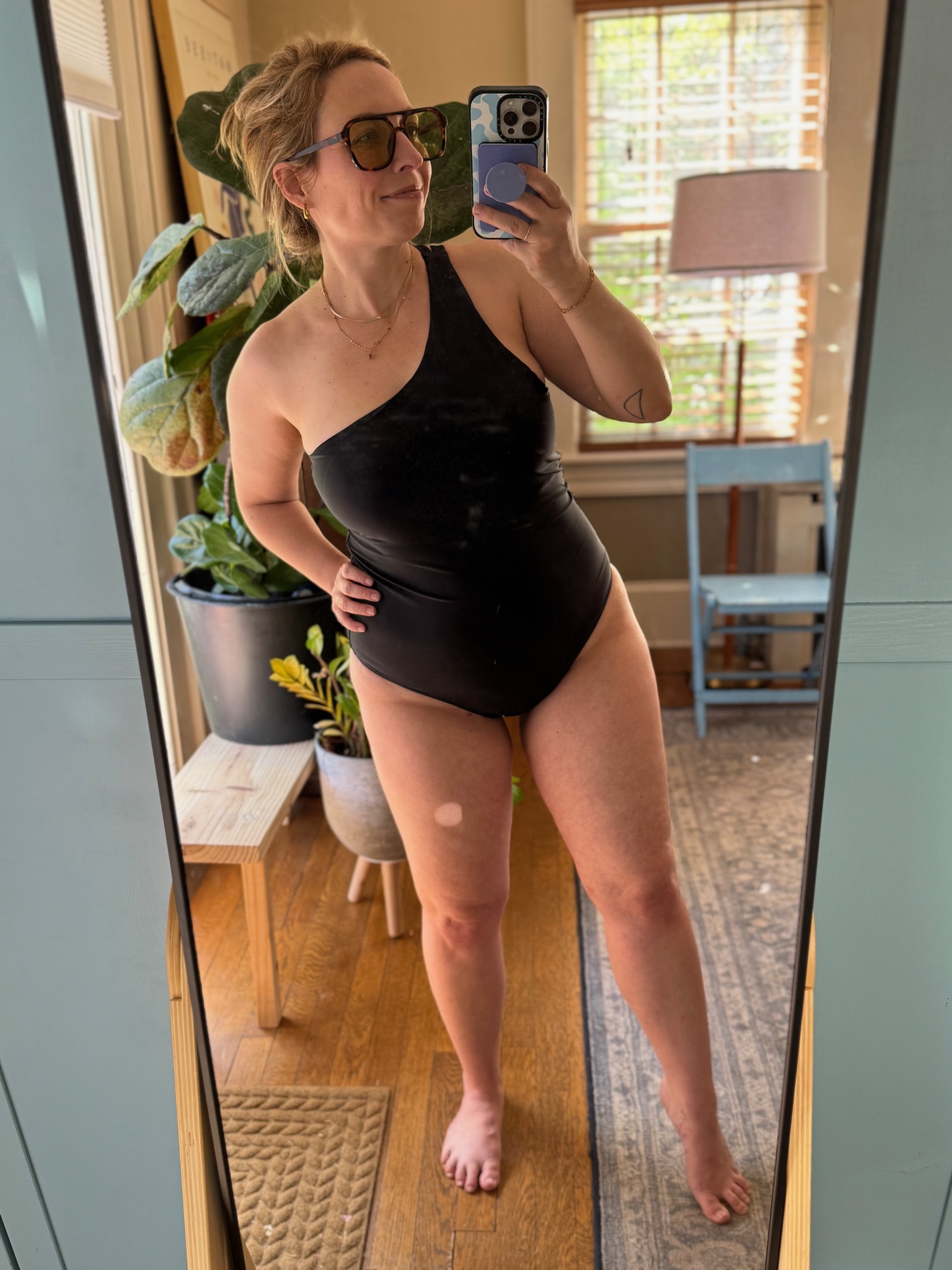 Woman wearing a black one-shoulder swimsuit takes a mirror selfie in a sunlit room with plants, a blue chair, and wooden floors visible in the background.